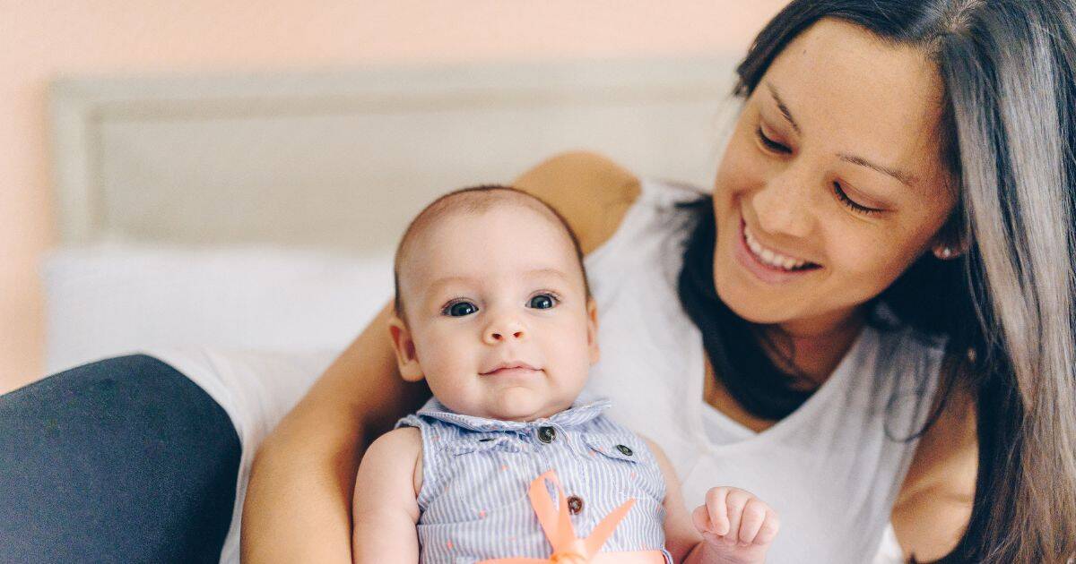 mommy and me portrait of a mother sitting on a bed behind her baby, she's looking and smiling at baby while baby shows a slight grin to the camera.