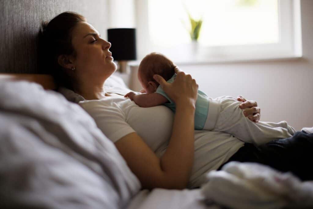 canva image of a new mom in bed looking exhausted while holding a newborn baby