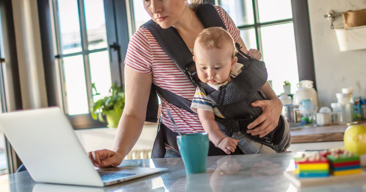 Image of a woman working on a laptop in a kitchen while holding a baby in a baby carrier.