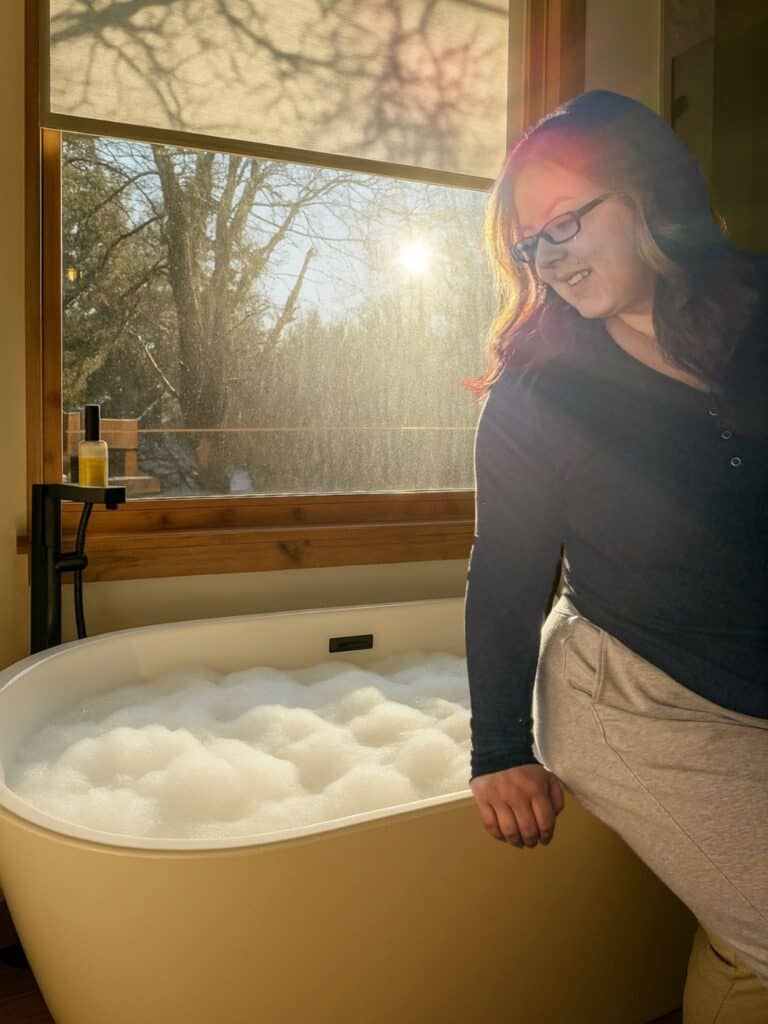 woman with a black top and grey bottoms leaning against a bath tub full of bubbles as the sun shines into the room