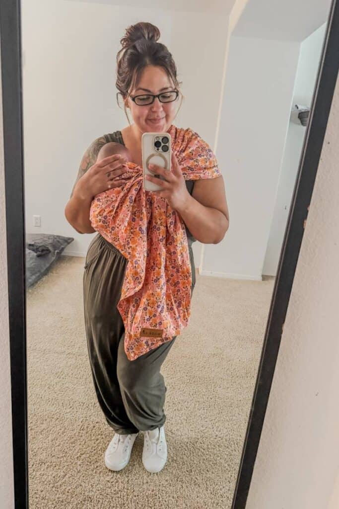 Karissa of Mom After Baby wearing a green romper with white sneakers and baby wearing with the Wildbird Sling Carrier.