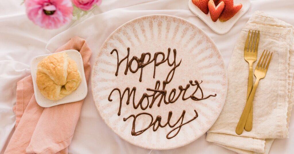 image of a breakfast spread with a croissant, some flowers and heart shaped strawberries next to a plate with chocolate writing that says 'happy mother's day'