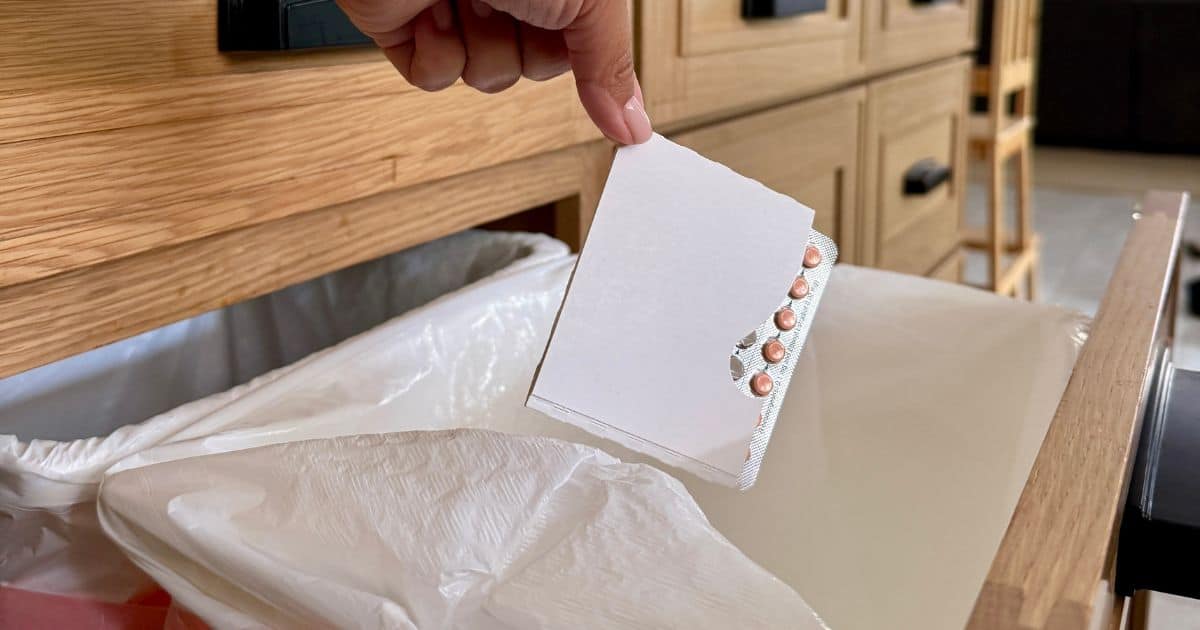 Image is a close up of a woman's hand throwing away a pack of birth control pills