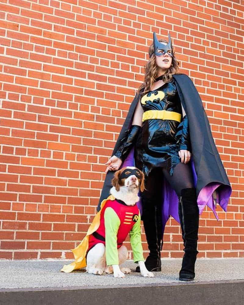 Image of a woman dressed as Batman with her dog dressed as Robin standing in front of a brick wall for Halloween.
