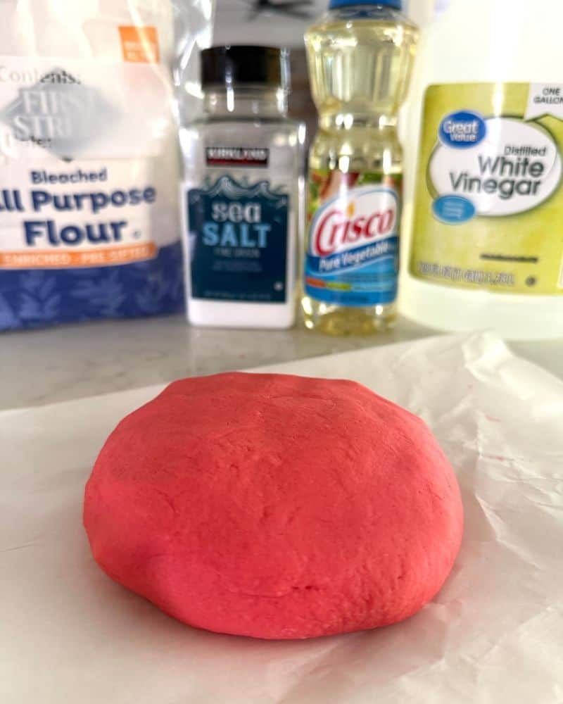 Homemade red playdough in front of flour, salt, oil, and vinegar ingredients.