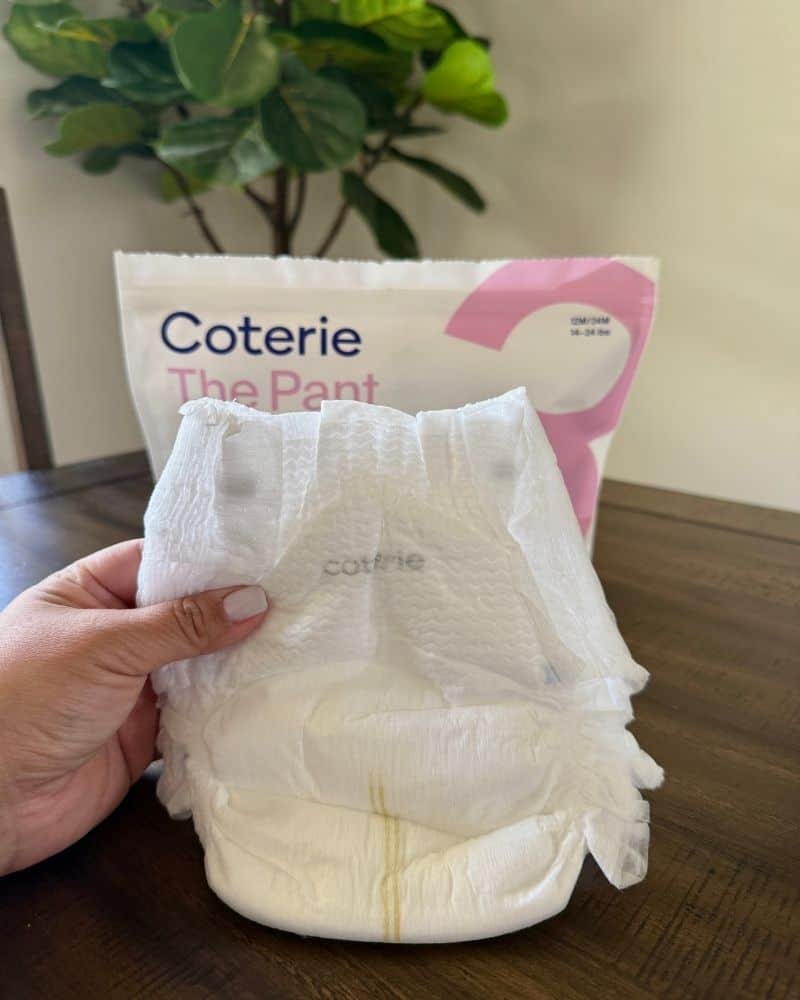 Image of a hand holding a Coterie diaper pant with the bag of Coterie diapers in the background
